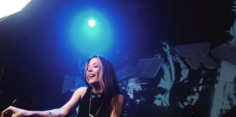Female DJ smiling under blue club lights with headphones around her neck during a DJ set in Dubai